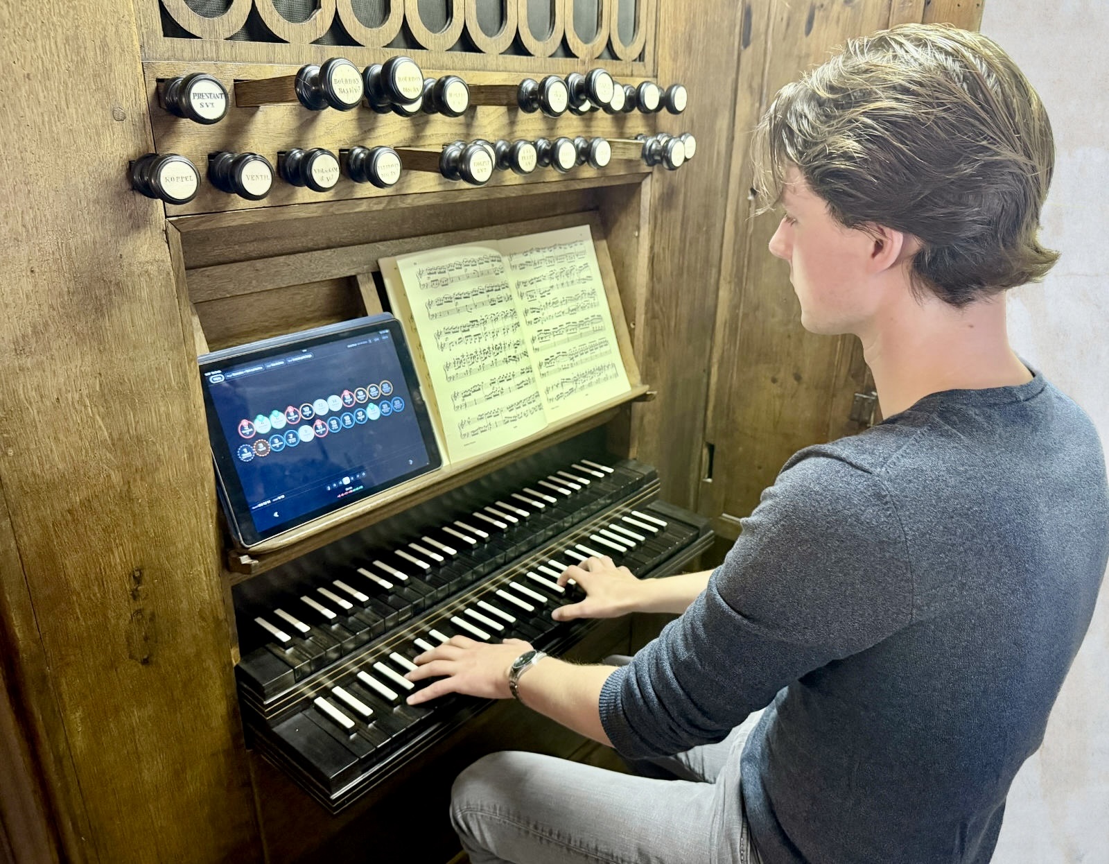 Een organist aan de speeltafel. Op de lessenaar staat een tablet met
                  Registrant: dezelfde registerknoppen als op het orgel, met de namen erin.
                  Daarboven de echte registerknoppen met kleine, moeilijk leesbare plaatjes.